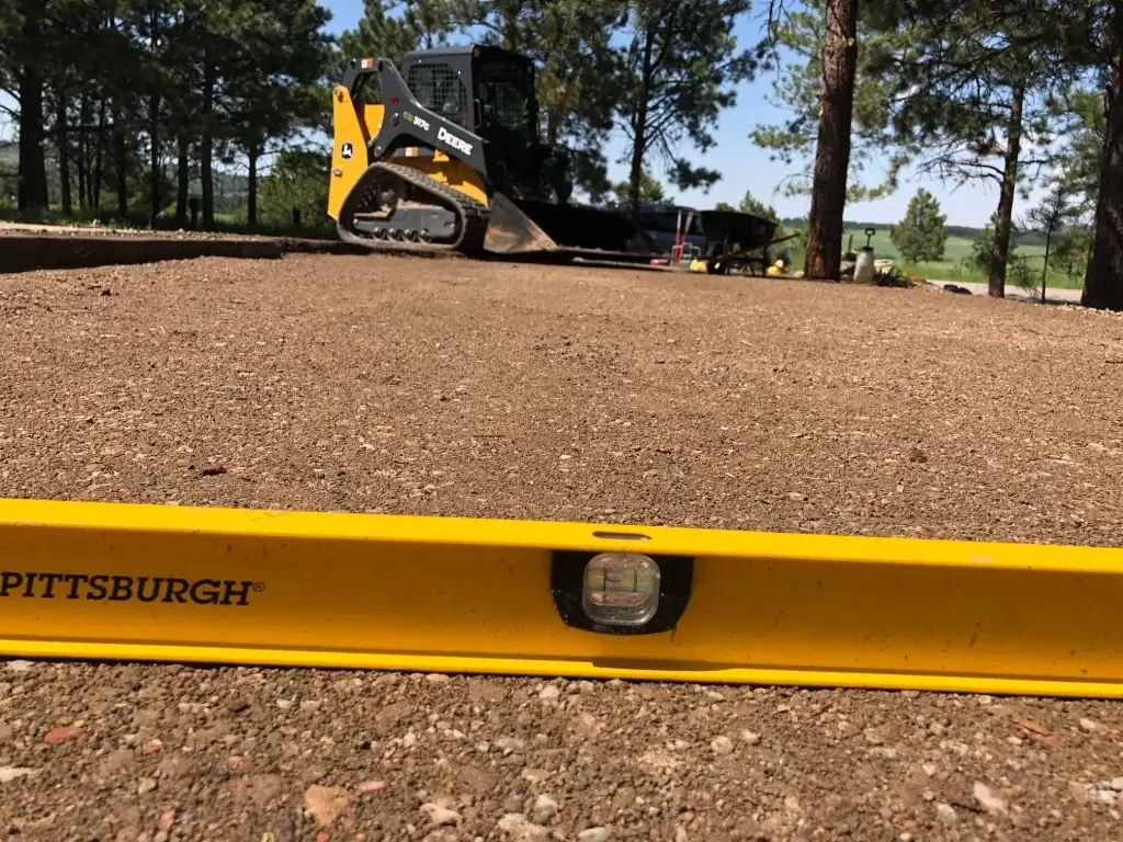 Skid steer and a level on a freshly graded gravel base under pine trees